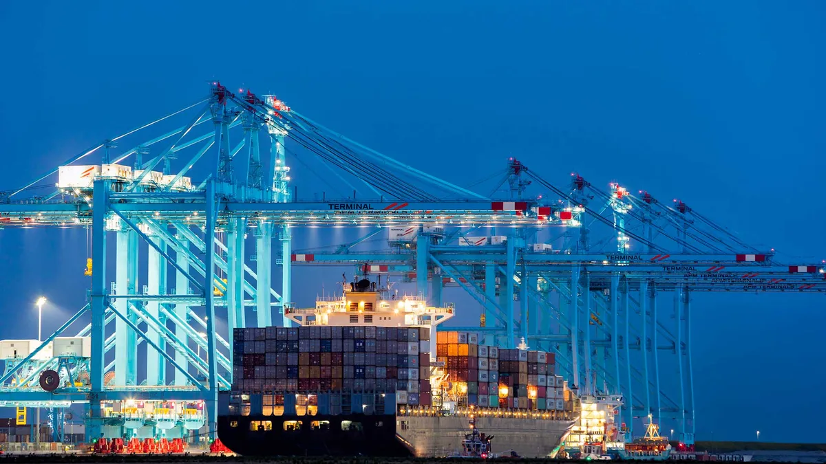 A container ship under gantry cranes at a port at dusk.
