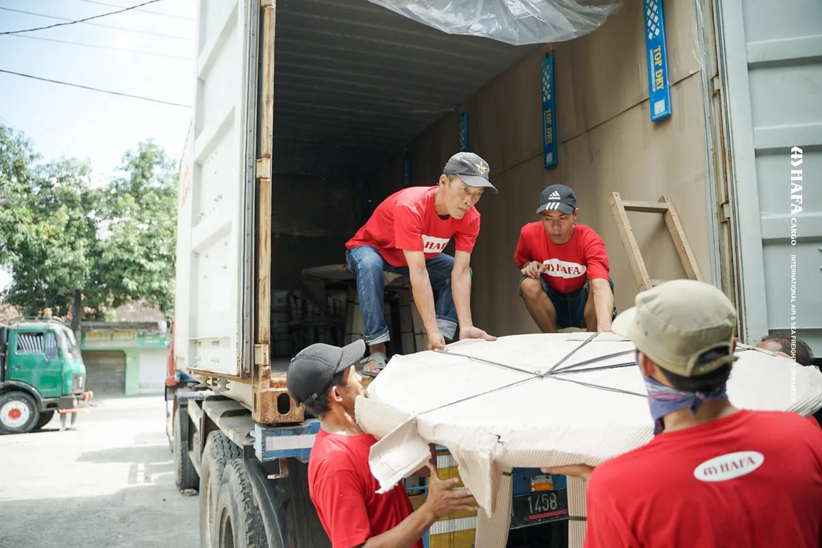 Tim HAFA Cargo mengangkat daun meja bundar besar yang sudah dibungkus ke dalam kontainer di atas truk.