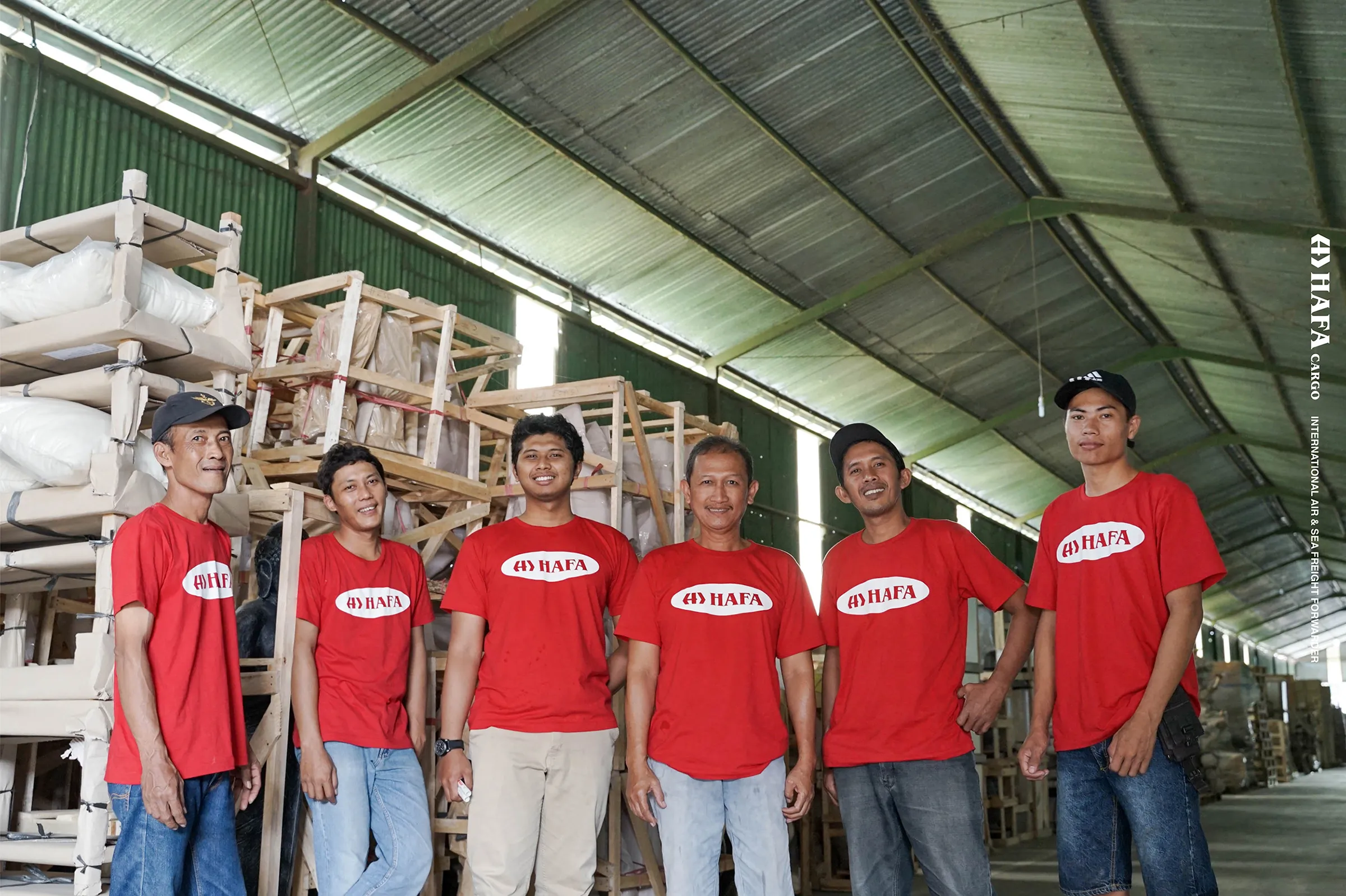 Seis miembros del equipo de HAFA Cargo con camisetas rojas de HAFA, en el almacén, delante de mercancía embalada en cajas de madera.