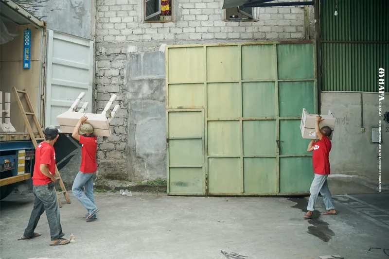 Tres empleados de HAFA Cargo cruzan el patio; dos de ellos llevan muebles embalados sobre la cabeza.